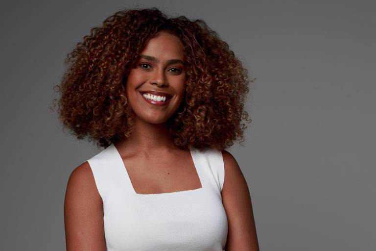 A imagem mostra uma mulher com cabelo cacheado e volumoso, usando uma blusa branca de alças largas. Ela está sorrindo e olhando diretamente para a câmera, com um fundo cinza neutro.