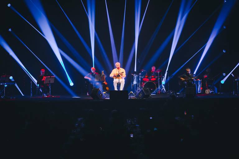 A imagem mostra um palco iluminado com feixes de luz azul e branca. No centro, um artista está se apresentando, vestido com uma camisa clara e calças brancas, segurando um instrumento musical. Ao seu redor, há quatro músicos tocando diferentes instrumentos, incluindo guitarra e percussão. O público é visível em primeiro plano, com algumas pessoas segurando celulares para gravar o show.