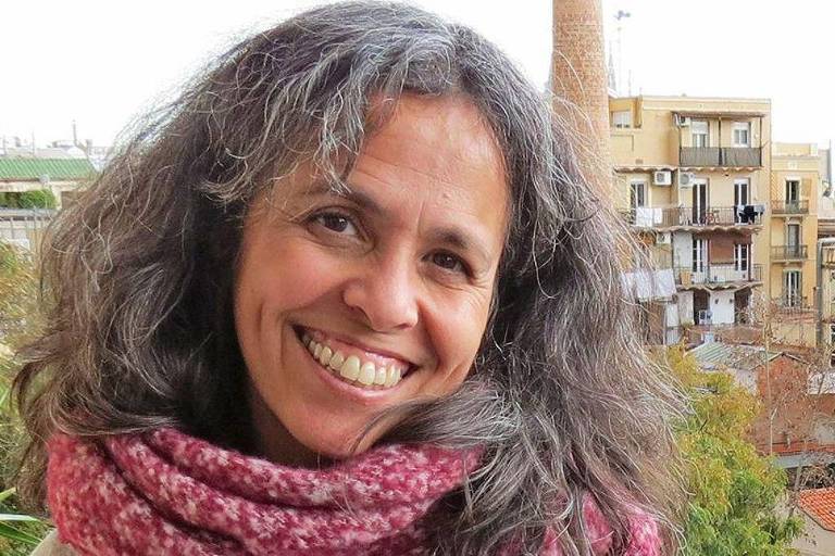 Uma mulher com cabelo cacheado e grisalho sorri para a câmera. Ela está usando um cachecol rosa e está em um ambiente externo， com prédios ao fundo. A cena parece ser em uma área urbana， com vegetação visível ao lado.