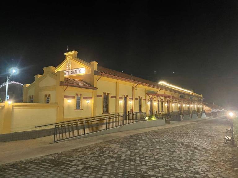 Imagem de uma estação de trem iluminada à noite. A estrutura é de cor amarela com detalhes em marrom. O nome 039;Estação039; está visível na parte superior. Há uma calçada em frente, com iluminação e um caminho acessível. O ambiente é tranquilo, com algumas luzes ao fundo.