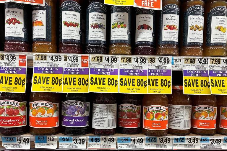 A imagem mostra uma prateleira de geleias em um supermercado. Há várias marcas e sabores， incluindo geleia de framboesa， damasco， uva， morango e outros. Os preços estão visíveis， com alguns produtos marcados com etiquetas de desconto， indicando 039;SAVE 80¢039;. Os preços variam de $3.49 a $7.98.