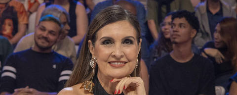 Domingão com Huck - Fátima Bernardes *** Local Caption *** Uma mulher vestida com elegância sorri confiante enquanto está sentada em um palco, com um copo de água e um smartphone à sua frente. Ela apoia o queixo na mão, exibindo unhas vermelhas que combinam com seu batom. Ao fundo, o público observa atentamente, sugerindo que ela pode ser uma palestrante ou jurada em um evento