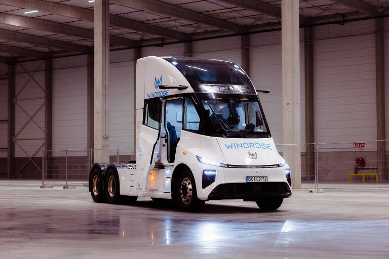 Um caminhão elétrico da marca Windrose, com um design moderno e aerodinâmico. O veículo possui uma cabine com janelas grandes e uma pintura branca com detalhes em azul. Está posicionado em um espaço amplo e vazio, com paredes de metal e piso de concreto.