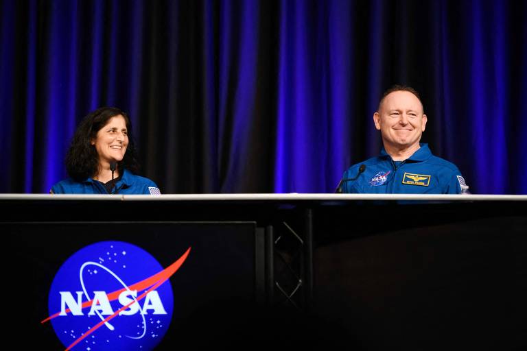 A imagem mostra dois astronautas em uma conferência da NASA. Eles estão sentados atrás de uma mesa, com um fundo de cortinas escuras e uma iluminação suave. O logotipo da NASA é visível na parte inferior da imagem, apresentando um círculo azul com estrelas e uma faixa vermelha.