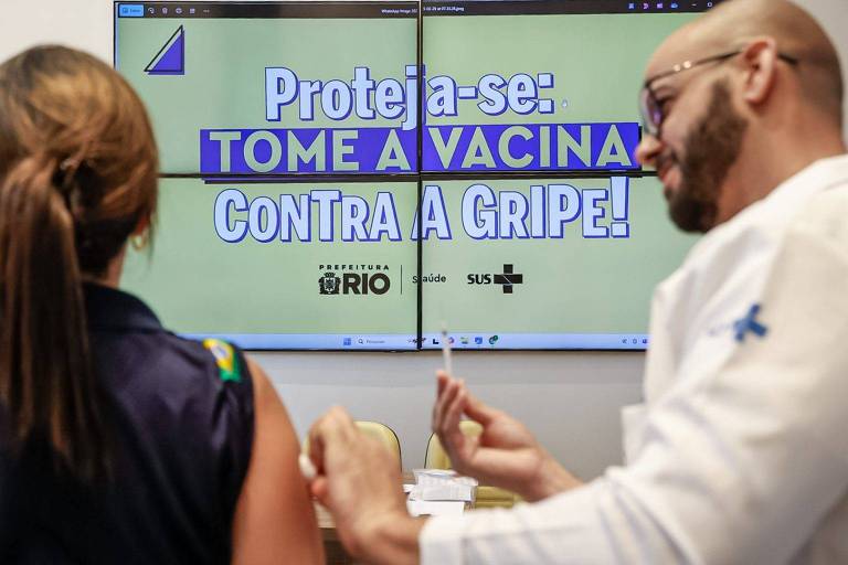 A imagem mostra uma tela com a mensagem 039;Proteja-se: TOME A VACINA CONTRA A GRIPE!039; em destaque. Ao lado， duas pessoas estão conversando， uma delas está de costas e a outra é um profissional de saúde， usando um jaleco e óculos. A tela também exibe logotipos da Prefeitura do Rio e do SUS.