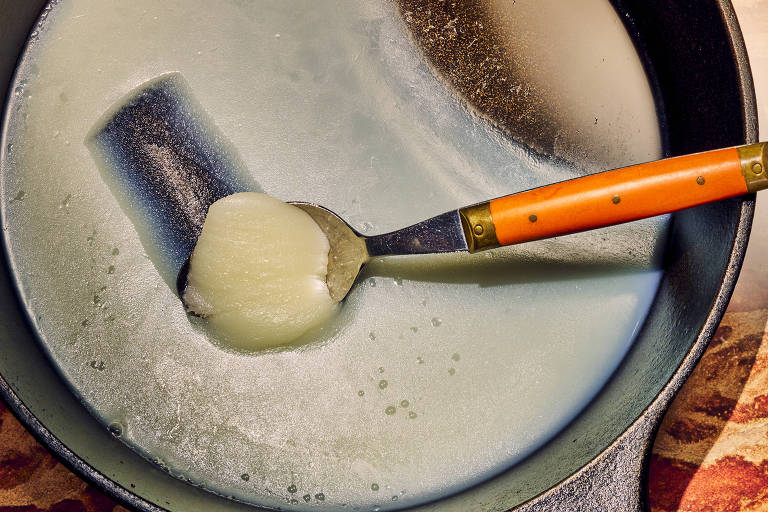 A imagem mostra uma panela de ferro fundido com uma camada de gordura derretida. Um utensílio de cozinha com cabo laranja está mergulhado na gordura， que é de cor clara e está em estado líquido. O fundo da imagem apresenta uma superfície com padrões em tons de marrom.