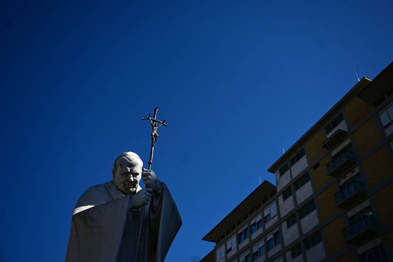 A imagem mostra uma estátua de um Papa segurando um crucifixo， com um céu azul claro ao fundo. À direita， há um edifício com várias janelas visíveis.