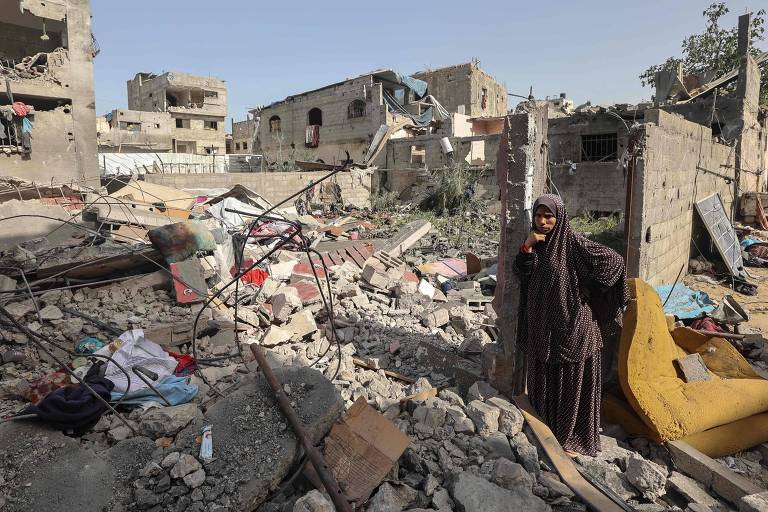 A imagem mostra uma mulher em pé entre os escombros de um edifício destruído. O cenário é de devastação, com prédios danificados ao fundo e destroços espalhados pelo chão. A mulher está vestida com uma roupa escura e parece estar observando a cena ao seu redor.