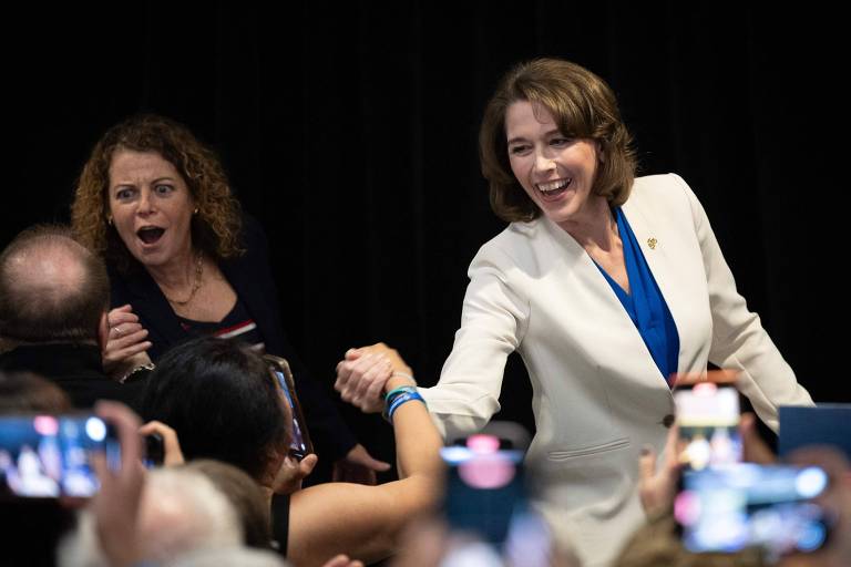 A imagem mostra uma mulher de blazer branco e blusa azul cumprimentando uma pessoa na plateia, enquanto outra mulher com cabelo cacheado e blusa listrada observa com expressão de surpresa. O ambiente parece ser um evento político, com várias pessoas ao fundo segurando celulares para registrar o momento.