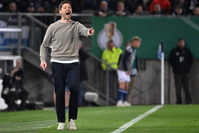 Um homem está em pé à beira do campo, vestindo um suéter cinza e calças escuras. Ele está gritando e gesticulando, demonstrando emoção durante um jogo de futebol. Ao fundo, há jogadores e um banner verde com um emblema. A atmosfera é de intensa atividade esportiva.