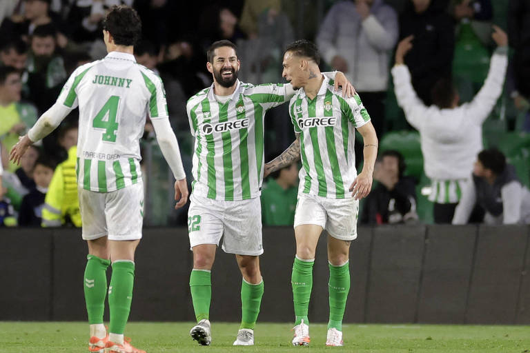 Três jogadores de futebol， que jogam pelo Betis， estão em um campo. O jogador à esquerda， identificado como 039;Johnny039;， usa uma camisa verde e branca com o número 4. O jogador do meio， com uma camisa semelhante， está sorrindo e tem o número 22. É Isco. O terceiro jogador， à direita， também veste uma camisa verde e branca e está sorrindo. É Antony. Ao fundo， há torcedores em um estádio.