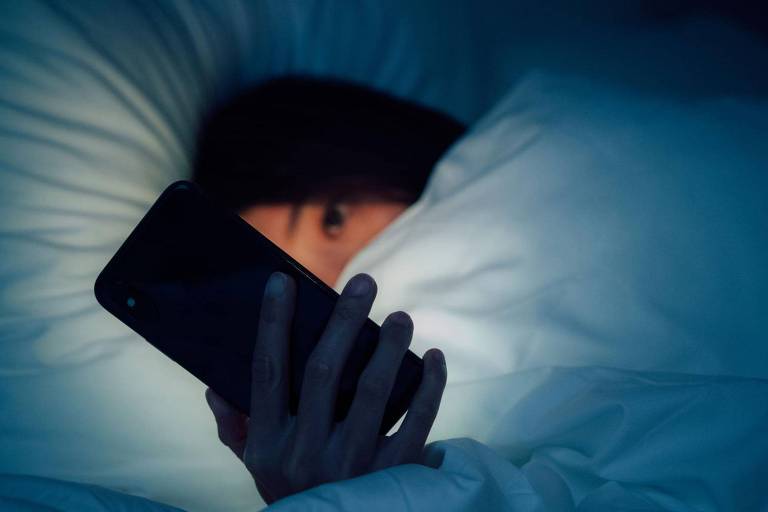 A imagem mostra uma pessoa deitada na cama, coberta com lençóis brancos, segurando um celular que emite uma luz suave. O ambiente é escuro, e a pessoa parece estar olhando para o celular, com apenas os olhos visíveis.