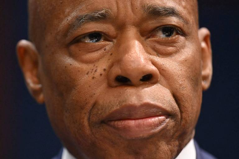 A imagem mostra um close de um homem com pele escura， expressando uma expressão séria. Ele tem cabelo raspado e está usando um terno escuro. O fundo é desfocado， destacando seu rosto.