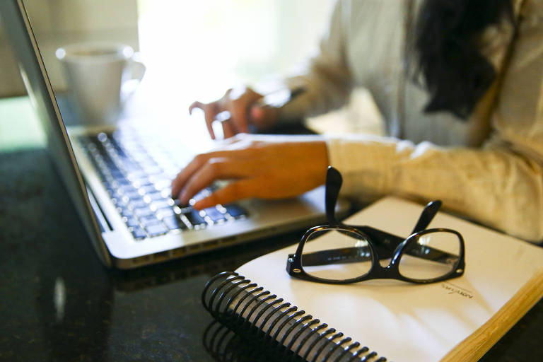 A imagem mostra uma pessoa digitando em um laptop. A mão da pessoa está sobre o teclado， enquanto a outra mão está visível ao lado. Em primeiro plano， há um caderno espiral e um par de óculos. Ao fundo， pode-se ver uma xícara.