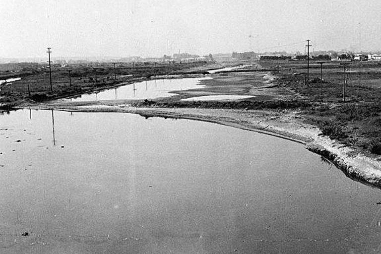 A imagem mostra um rio curvado em uma área rural， com água refletindo o céu. À margem do rio， há uma faixa de terra e vegetação escassa. No fundo， podem ser vistos postes de eletricidade e uma área urbana ao longe， com edifícios e estruturas. A imagem é em preto e branco， sugerindo um estilo vintage.