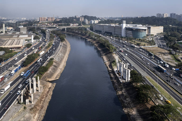 Imagem aérea mostrando uma estrada movimentada com veículos em ambos os sentidos， ao lado de um rio. À direita， há um edifício moderno e， à esquerda， estruturas de concreto que parecem ser pilares. O céu está claro e a vegetação é visível nas margens do rio.