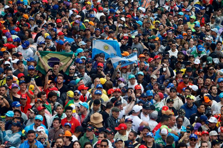 A imagem mostra uma grande multidão reunida em um evento， com pessoas usando bonés de várias cores. Algumas bandeiras são visíveis， incluindo a bandeira da Argentina e do Brasil. A multidão é densa e as pessoas parecem estar animadas， com muitas delas segurando celulares