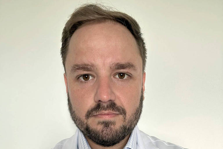 Um homem com barba e cabelo curto, vestindo um jaleco branco. Ele está olhando diretamente para a câmera, com uma expressão séria. O jaleco possui um nome bordado que diz 'Dr. Elmar D.' e a função 'Diretor Médico'. O fundo é claro e neutro.