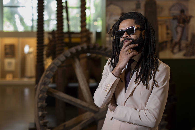 Um homem com cabelo longo e dreadlocks， usando óculos escuros e um terno claro， está posando com uma mão no queixo em um ambiente de museu. Ao fundo， há uma grande roda de madeira e várias exposições visíveis nas paredes.