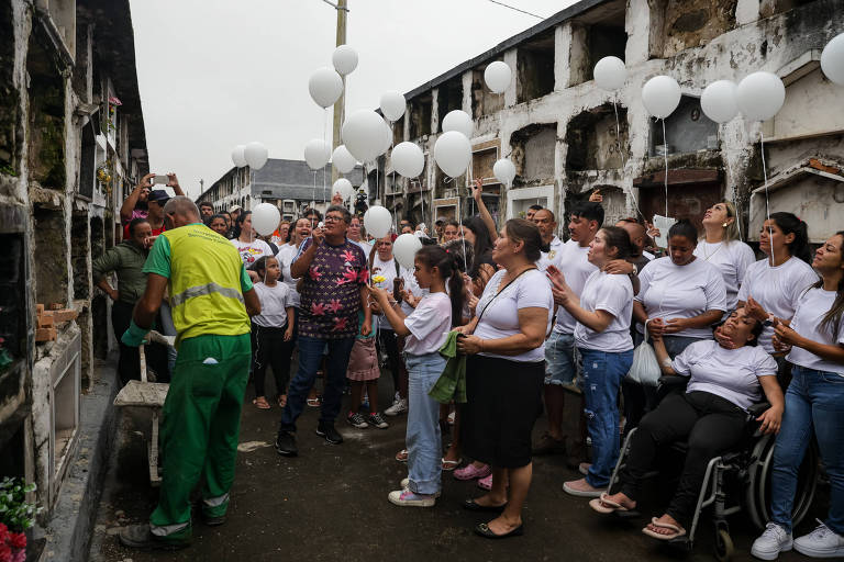 A imagem mostra um grupo de pessoas em um cemitério， algumas segurando balões brancos. Há um homem vestido com uniforme de trabalho， que parece estar realizando uma atividade relacionada ao local. As pessoas estão vestidas principalmente com camisetas brancas e algumas estão em uma cadeira de rodas. O ambiente é sombrio， com túmulos visíveis ao fundo e um céu nublado.