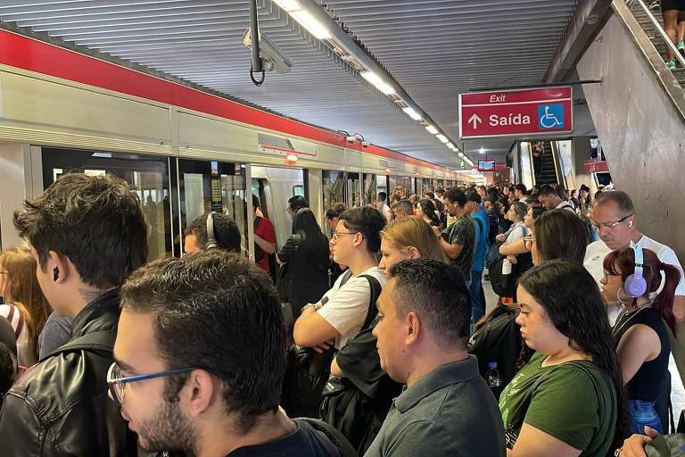 A imagem mostra uma estação de metrô lotada， com muitas pessoas aguardando na plataforma. Há um trem visível à esquerda， e um sinal indicando a saída à direita. A maioria das pessoas está de pé， algumas usando fones de ouvido， enquanto outras olham para o trem.