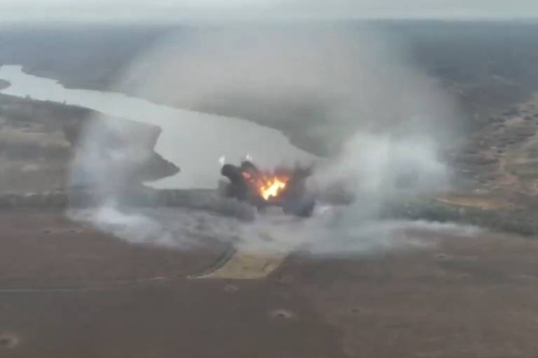 A imagem mostra uma explosão em uma área rural， com fumaça e chamas visíveis. Ao fundo， há um rio serpenteando pela paisagem e uma vegetação variada. O céu está nublado， e a cena é capturada de uma perspectiva aérea.