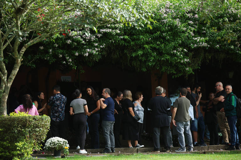 A imagem mostra um grupo de pessoas em uma fila， em um ambiente ao ar livre， sob uma árvore com folhagem densa. Algumas pessoas estão conversando， enquanto outras parecem esperar. O cenário é tranquilo， com um gramado verde e flores brancas visíveis em primeiro plano.