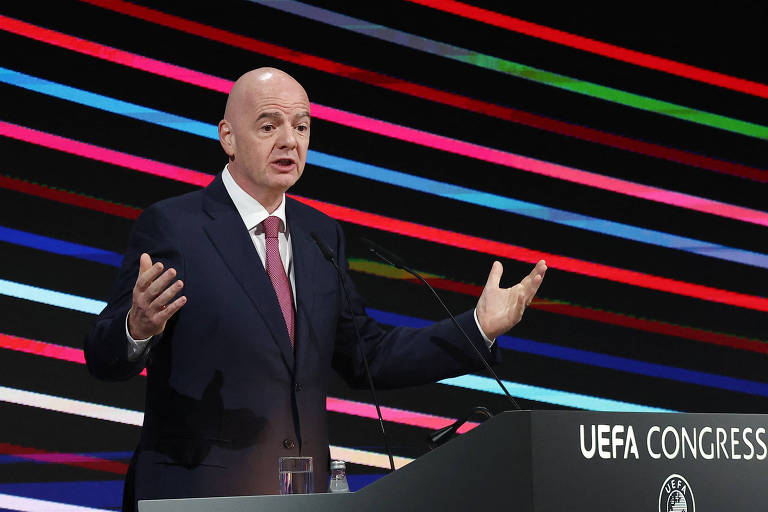 Um homem de cabeça calva， vestido com um terno escuro e gravata， está em pé atrás de um púlpito com o logotipo da UEFA. Ele gesticula com as mãos enquanto fala， e ao fundo há linhas coloridas em um padrão horizontal.