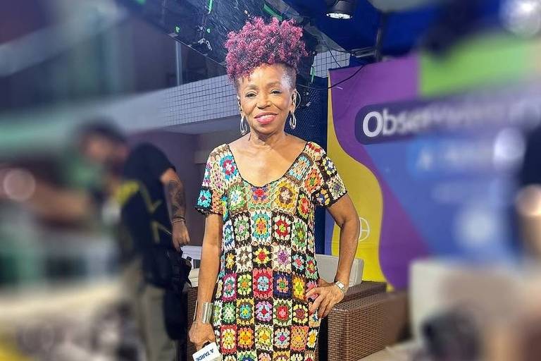 Uma mulher está em pé, sorrindo, usando um vestido longo com estampas coloridas em formato de flores. Ela tem cabelo crespo e volumoso, tingido de rosa. O fundo apresenta um ambiente de estúdio com sofás e uma parede colorida com o texto 039;Observatório039;.