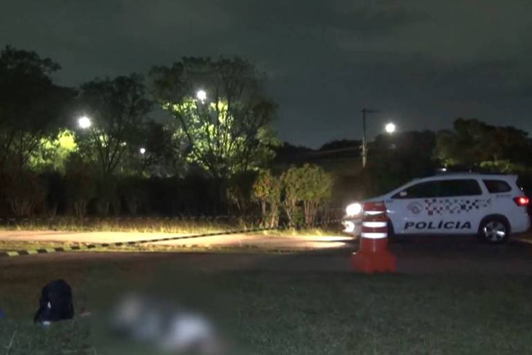 A imagem mostra uma cena de crime à noite， com um veículo da polícia estacionado em uma área iluminada por postes de luz. Há cones de sinalização laranja e uma fita amarela delimitando a área. Ao fundo， é possível ver árvores e um espaço aberto. No chão， há uma mancha que não é claramente identificável.