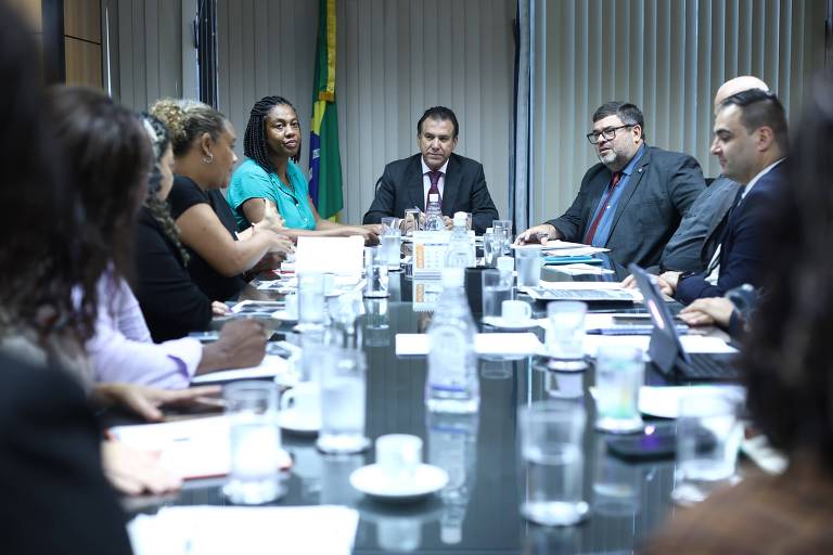 A imagem mostra uma reunião em uma mesa redonda com várias pessoas sentadas. Há um total de oito pessoas visíveis, incluindo homens e mulheres, discutindo documentos e utilizando laptops. Ao fundo, há uma bandeira do Brasil. A mesa está coberta com copos de água, xícaras e papéis.