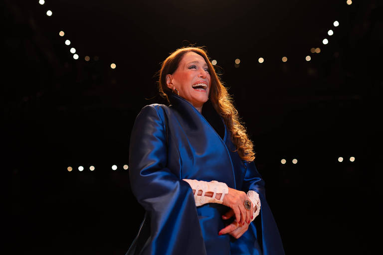 Uma mulher com cabelo longo e solto, vestindo um traje azul escuro, está sorrindo enquanto se encontra em um palco iluminado. O fundo é escuro, com luzes brilhantes visíveis, criando um ambiente de espetáculo.