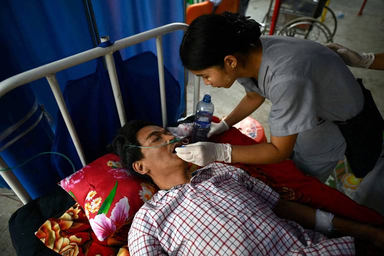 A imagem mostra um homem deitado em uma cama hospitalar, recebendo cuidados de uma profissional de saúde. O homem está com um tubo de oxigênio no rosto e parece estar inconsciente. A profissional, vestindo um uniforme cinza e luvas, está inclinada sobre ele, segurando uma garrafa de água e um cotonete. O fundo é composto por cortinas azuis e outros equipamentos médicos.