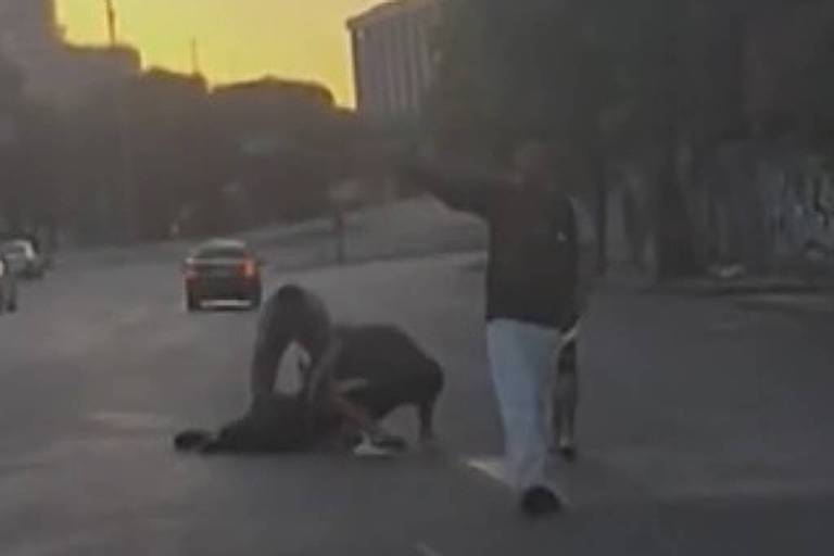 A imagem mostra duas pessoas em uma rua. Uma delas está deitada no chão, enquanto a outra está em pé, aparentemente interagindo com a pessoa no chão. O cenário é urbano, com edifícios ao fundo e uma luz suave do pôr do sol. Não há veículos visíveis na rua.