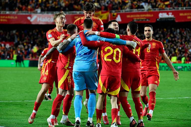 Um grupo de jogadores da seleção espanhola de futebol celebra em campo após um gol. Eles estão vestidos com uniformes vermelhos e amarelos, e alguns jogadores estão se abraçando enquanto outros estão em posições de comemoração. O estádio ao fundo está cheio de torcedores.