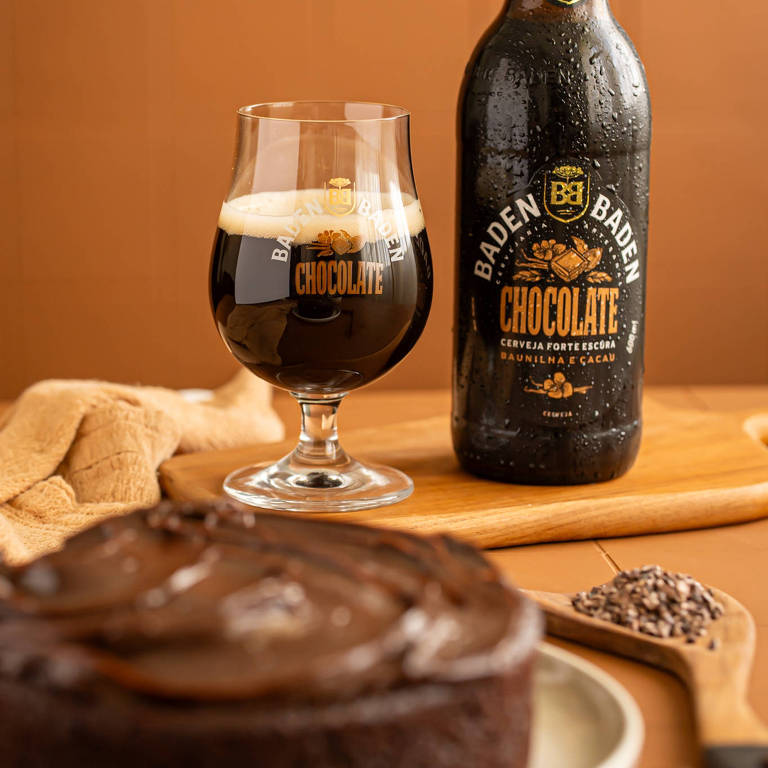 A imagem mostra uma garrafa de cerveja Baden Baden Chocolate ao lado de um copo com a mesma bebida， que apresenta uma cor escura e espumosa. Em primeiro plano， há um bolo de chocolate coberto com um glacê brilhante. O fundo é de uma cor neutra， e há uma colher com grãos de cacau ao lado do bolo.