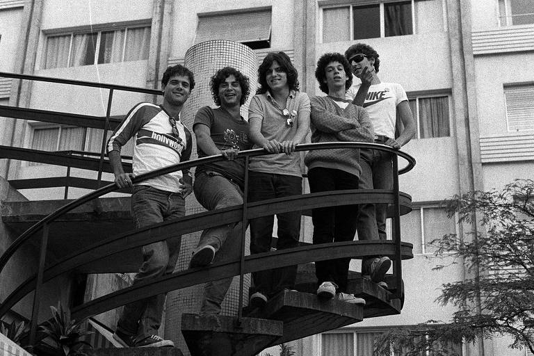 Cinco pessoas posando em uma escada em espiral, com um edifício ao fundo. Eles estão sorrindo e se divertindo. O ambiente é urbano, com janelas visíveis no prédio. As pessoas estão vestindo camisetas e jeans, e a imagem é em preto e branco.