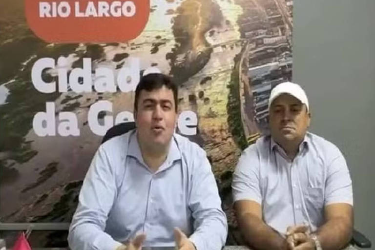 Imagem mostra dois homens sentados, gesticulando para um vídeo. Eles vestem camisas claras e um deles está de boné.