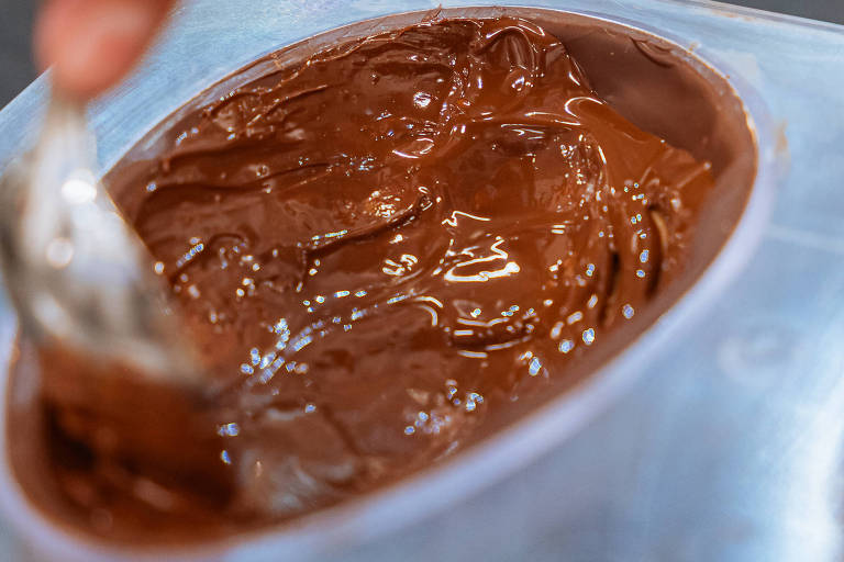 A imagem mostra uma mão segurando uma colher, mexendo uma mistura de chocolate em um recipiente oval. O chocolate tem uma textura cremosa e brilhante, e o fundo é uma superfície escura.