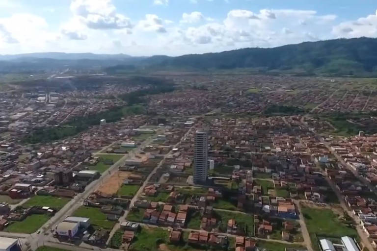 A imagem mostra uma vista aérea de uma cidade com uma grande extensão de áreas urbanas e rurais. Há uma mistura de edifícios baixos e uma torre alta em destaque. O fundo é composto por montanhas e um céu parcialmente nublado.