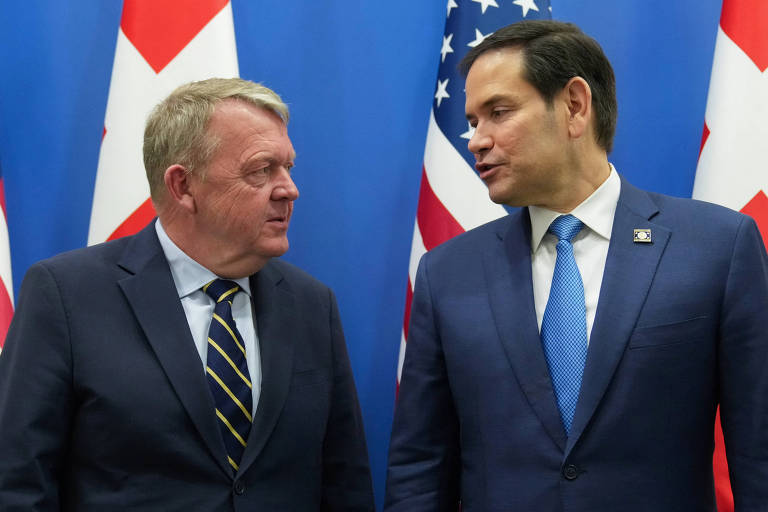 A imagem mostra dois homens em um encontro, conversando um com o outro. O homem à esquerda tem cabelo loiro e usa um terno escuro com uma gravata listrada. O homem à direita tem cabelo castanho e usa um terno escuro com uma gravata azul. Ao fundo, há bandeiras dos Estados Unidos e de outro país, com predominância de vermelho e branco.