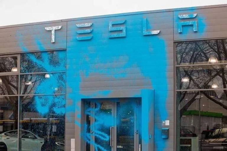 A imagem mostra a fachada de uma loja Tesla, onde o nome 039;TESLA039; está visível na parte superior. A parede de madeira está coberta com tinta azul, que parece ter sido aplicada de forma irregular, criando um efeito de vandalismo. A entrada da loja é de vidro, permitindo ver o interior, onde há um carro branco estacionado. O céu está nublado, e há árvores visíveis ao fundo.