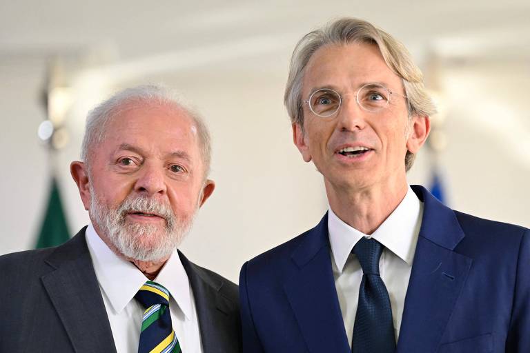 A imagem mostra dois homens sorrindo em um evento. O homem à esquerda tem cabelo grisalho e uma barba curta, vestindo um terno escuro com uma gravata listrada em verde e amarelo. O homem à direita tem cabelo loiro e usa óculos, vestido com um terno azul escuro e uma gravata preta. Ao fundo, são visíveis bandeiras.