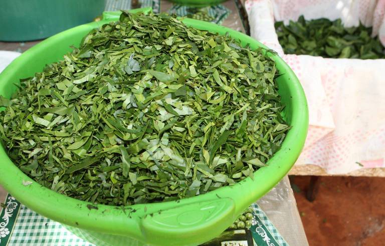 Uma tigela verde cheia de folhas verdes picadas, com uma textura fina e uniforme. Ao fundo, há uma mesa com mais folhas em um recipiente branco. O ambiente parece ser um mercado ou feira.