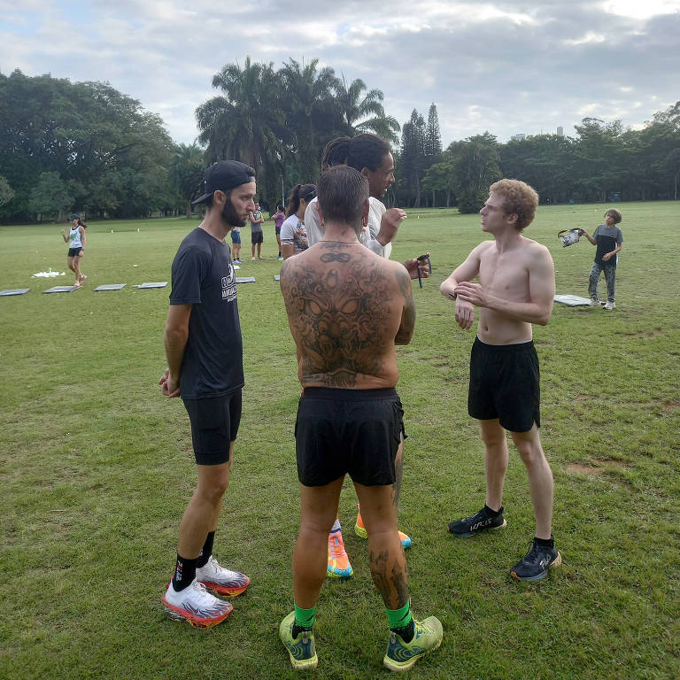 Quatro homens estão conversando em um campo gramado. Dois deles estão de camiseta e shorts, enquanto um está sem camisa e o outro usa uma camiseta preta. Ao fundo, outras pessoas estão visíveis, algumas em pé e outras deitadas em colchonetes. O céu está nublado e há árvores ao redor.