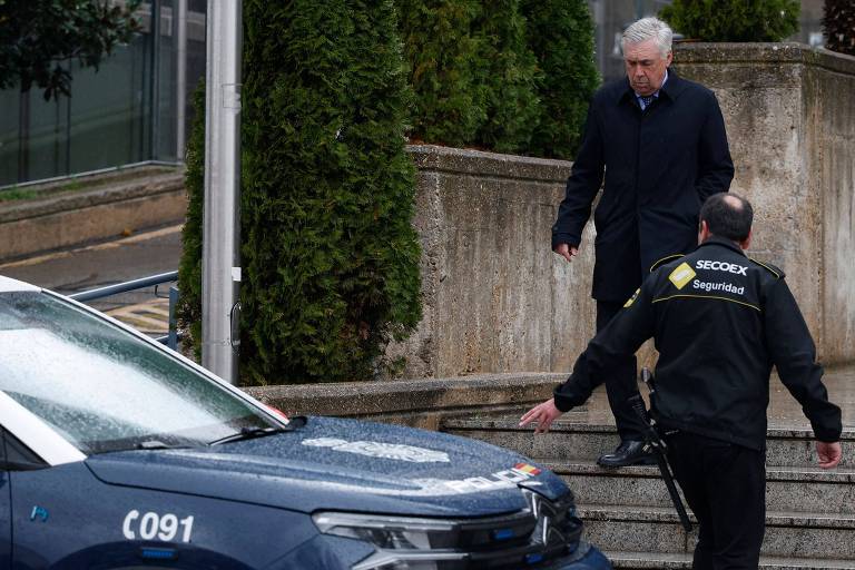 Um homem de cabelo grisalho está descendo uma escada em direção a um carro da polícia, enquanto um policial em uniforme preto se aproxima dele. O ambiente é urbano, com vegetação ao fundo e um dia nublado.
