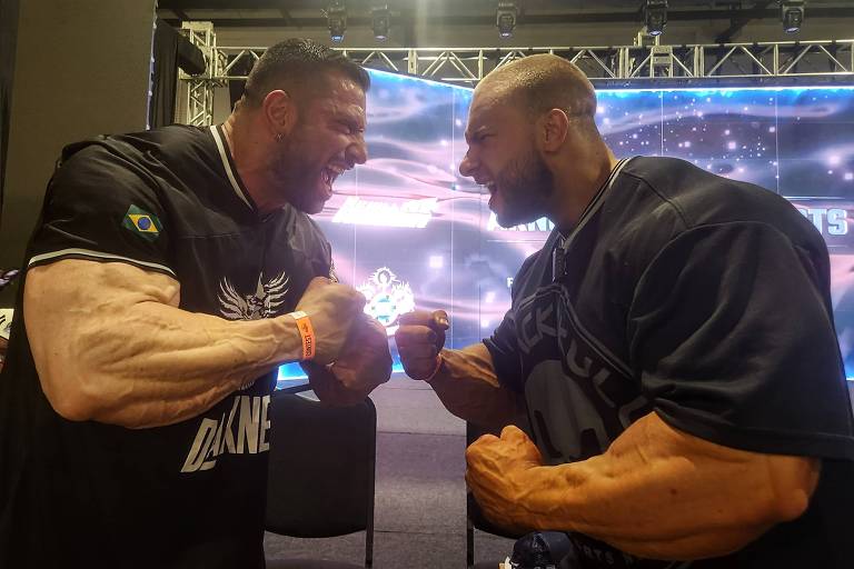 Dois fisiculturistas estão se enfrentando em uma competição, mostrando seus músculos e fazendo poses de força. Ambos têm braços muito musculosos e estão com expressões faciais intensas. O fundo apresenta uma tela iluminada com luzes, sugerindo um ambiente de evento esportivo.