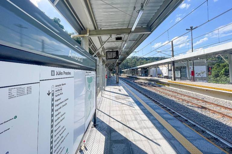 A imagem mostra uma plataforma de estação de trem com um teto coberto. À esquerda, há um painel informativo com uma lista de destinos. A plataforma é ampla e bem iluminada, com trilhos visíveis ao fundo. O céu está claro e azul, e há algumas nuvens. Não há pessoas visíveis na imagem.