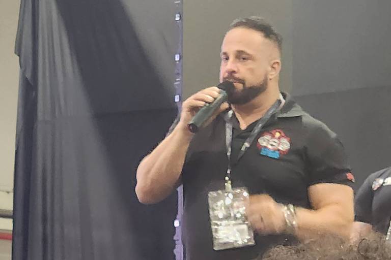 A imagem mostra um homem em pé, segurando um microfone e falando. Ele tem cabelo curto e barba, vestindo uma camisa preta com um logotipo. Ao fundo, há outro homem com cabelo curto, também vestido de preto. O ambiente parece ser um evento, com cadeiras pretas visíveis e um fundo escuro.