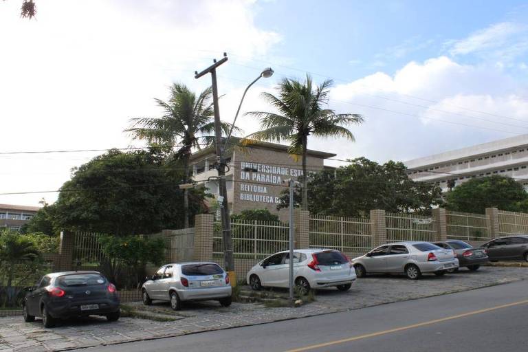 A imagem mostra uma instituição de ensino com um edifício de dois andares ao fundo, cercado por árvores e palmeiras. Na frente, há um estacionamento com vários carros estacionados. O céu está parcialmente nublado.